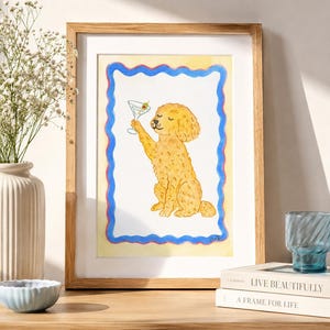 Könnte beinhalten: Ein gerahmter Aquarelldruck, der einen goldenen, lockigen Hund zeigt, der sitzt und ein Martiniglas mit einer Olive hält. Das Kunstwerk ist von einer wellenförmigen blauen Linie auf hellgelbem Hintergrund umrandet und in einem hellen Holzrahmen.