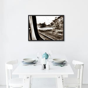 Può includere: Una fotografia incorniciata in bianco e nero appesa a una parete sopra un tavolo da pranzo bianco. L'immagine raffigura il finestrino di un treno coperto di gocce di pioggia, che si affaccia su un paesaggio sfocato con binari in toni seppia.