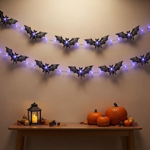May include: Two strings of black bat-shaped lights with glowing purple LEDs are hung horizontally. Below, a black metal lantern with a lit candle sits on a table next to three orange pumpkins, a small gourd, and scattered brown pinecones and leaves.