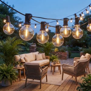 May include: A string of outdoor globe lights with clear glass bulbs and black sockets suspended on a dark wire. The bulbs emit a warm, glowing light, illuminating a patio area featuring a beige sofa, two woven chairs, and small wooden tables.