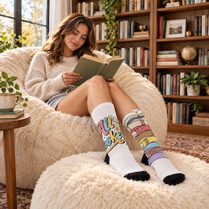 May include: A pair of white crew socks featuring a graphic design. The left sock displays the text "All Books" in a colorful, stylized font with small star accents. The right sock features a vertical stack of multi-colored books with black toes.