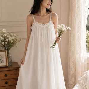 May include: A white sleeveless nightdress featuring a gathered bodice, thin adjustable shoulder straps, and a delicate ruffled trim along the neckline. The garment is crafted from a lightweight, textured fabric that falls into a loose, flowing skirt.
