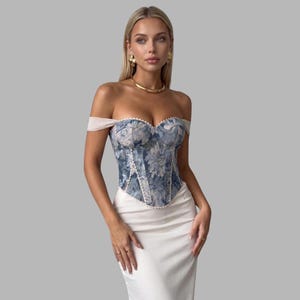 May include: An off-the-shoulder corset top featuring a blue and white floral pattern with white trim detailing, paired with a plain white skirt. The corset has a sweetheart neckline and structured boning, complemented by gold hoop earrings and a gold necklace.
