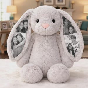 Op de afbeelding: Een lichtgrijze pluche konijnenknuffel met lange, slappe oren. Op elk oor is een zwart-wit fotocollage van mensen afgebeeld. Het konijn heeft een zachte, pluizige textuur, ronde zwarte ogen en een kleine, lichtroze neus.