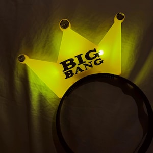 May include: A yellow illuminated crown-shaped headband featuring the text BIG BANG in black lettering. The crown has three points, each topped with a small circular accent, and is attached to a thin, black, semi-circular plastic headband frame.