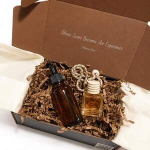May include: A brown cardboard gift box containing an amber glass dropper bottle and a small square glass bottle with a wooden cap and hanging rope. The inside lid features the text Where Scent Becomes An Experience and Thank You! nestled in brown paper.