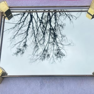 May include: A rectangular vanity tray featuring a reflective mirror base and a thin metal frame. The tray is accented with four decorative gold-toned metal corner pieces, each displaying a fan-like shape with distinct ridges at the edges.