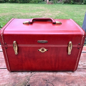 May include: A rectangular red Samsonite hard-shell travel case featuring a top handle, gold-toned metal latches, and a central gold-toned keyhole lock. The exterior has visible white stitching along the edges and a small metal brand logo plate.