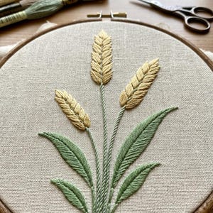 Pode incluir: Um bastidor de bordado contendo um painel de tecido bege com um desenho costurado à mão de três espigas de trigo em fio amarelo e várias folhas verdes. O bordado é rodeado por três meadas de fio de bordar e tesouras de metal.