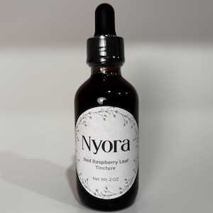 May include: A dark amber glass dropper bottle with a black cap and a white oval label. The label features a floral border and black text reading Nyora, Red Raspberry Leaf, Tincture, and Net Wt. 2 OZ. The bottle contains a dark liquid.