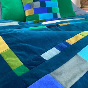 May include: A teal velvet quilt with a patchwork design featuring squares and rectangles in shades of blue, green, and gold. Two solid green velvet pillows and a patterned pillow with a similar color scheme complete the bedding set.