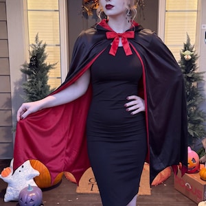 May include: A reversible hooded cape featuring a smooth black outer layer and a deep red inner lining. The cape is secured at the neck with a matching red satin ribbon bow tie. The hem of the cape is cut into distinct, pointed bat-wing shapes.