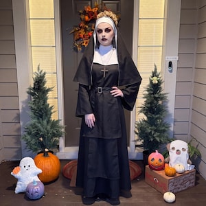 May include: A black and white nun costume featuring a long-sleeved black habit, a white wimple, a black veil, and a black belt with a silver buckle. A silver-toned cross pendant hangs from a chain around the neck, completing the traditional look.