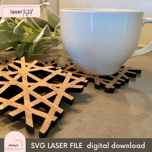 Puede incluir: Dos posavasos de madera con forma de copo de nieve y un contorno negro. Los posavasos son de madera marrón claro y están sobre una superficie marrón claro. Una taza de café blanca está encima de los posavasos.