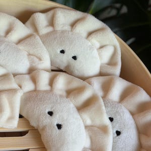 Könnte beinhalten: Eine Sammlung weicher, cremefarbener Plüsch-Teigtaschen mit gefalteten Rändern und kleinen, gestickten schwarzen Augen. Jede Plüsch-Teigtasche hat eine halbkreisförmige Form und eine flauschige Textur und ist in einem hellen Holzdampfgarer angeordnet.