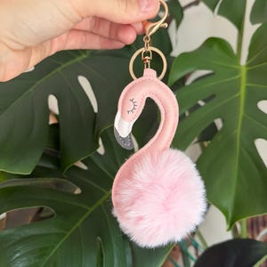 May include: A flamingo-shaped keyring featuring a smooth pink leatherette neck and head with a white and grey beak, accented by a soft, fluffy light pink faux fur pom-pom body. The accessory includes a gold-toned metal lobster clasp and ring.