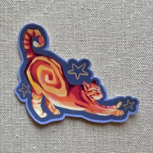 May include: A die-cut sticker featuring an orange tabby cat in a stretching pose with a spiral pattern on its side. The cat is surrounded by three small, outlined stars against a dark blue border, all set against a textured grey fabric background.