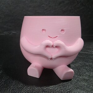 May include: A light pink planter shaped like a rounded character with two arms forming a heart shape in front of its chest. The character has a simple engraved face with two small eyes and a curved mouth, and two small feet at the base.
