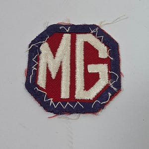 May include: An octagonal embroidered patch featuring the white letters MG centered on a red background. The patch is bordered by a thick dark blue edge with visible white stitching along the perimeter and loose threads at the corners.