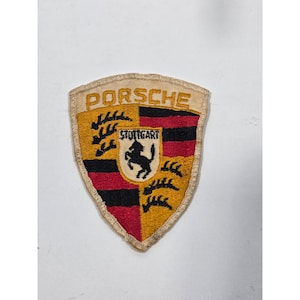 May include: A shield-shaped embroidered patch featuring the Porsche logo in gold, black, and red. The design includes the word PORSCHE at the top, a central crest with a black horse, and the text STUTTGART, surrounded by black and red stripes.