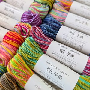 May include: A collection of multi-colored embroidery thread skeins arranged in a grid. Each skein features a white paper band labeled HOBBYRA HOBBYRE with Japanese text, specifying 100% cotton, 10g weight, and approximately 56m length.