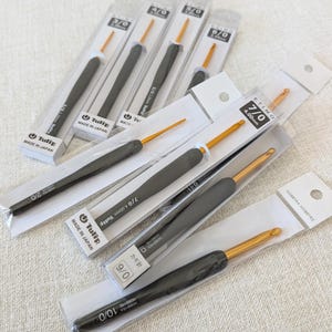 May include: A collection of Tulip Etimo and Hobbyra Hobbyre crochet hooks in individual clear plastic packaging. Each hook features a grey ergonomic handle and a gold-colored metal hook tip. The packaging displays size labels such as 3/0, 4/0, 5/0, 6/0, 7/0, 9/0, and 10/0, with text indicating Made in Japan and specific brand names printed on the white backing cards.
