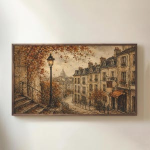 Pode incluir: Uma impressão de arte de parede retangular emoldurada retratando uma cena de rua europeia no outono. A imagem apresenta um poste de luz preto alto, escadas de pedra e edifícios de vários andares com folhagem marrom e laranja sob um céu.