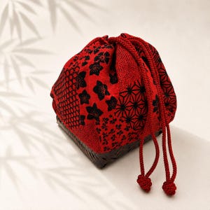 May include: A drawstring pouch featuring a vibrant red fabric with black floral and geometric patterns. The pouch is secured with two thick red braided cords that hang down with knotted ends. The base of the pouch is a dark brown woven basket.