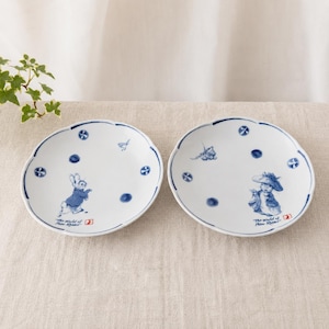 Puede incluir: Dos platos de cerámica blanca con bordes festoneados, decorados con ilustraciones azules de personajes de Peter Rabbit y pequeños motivos florales. Cada plato presenta el texto The World of Peter Rabbit y un pequeño logotipo rojo.