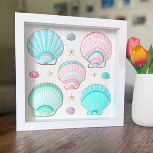 May include: A square white shadow box frame containing five large scallop shells painted in pastel shades of turquoise, pink, and mint green with gold-painted edges, interspersed with small starfish and tiny decorative shell accents.