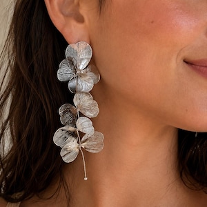 May include: A long silver-toned earring featuring a vertical arrangement of five delicate, filigree-style flower petals. The metal petals have a fine, linear texture and are connected by thin silver chains, ending in a small, singular metal bead.