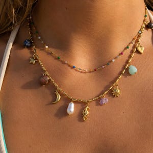 Könnte beinhalten: Ein mehrlagiges goldfarbenes Halskettenset bestehend aus einer feinen Kette mit kleinen bunten Perlen und einer dickeren Kette mit zylindrischen Gliedern. Die Anhänger umfassen einen Stern, Mond, ein Seepferdchen, Herz, Sonne, eine weiße Perle und Steine.