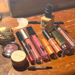 May include: A collection of makeup products including a Nude by Nature powder container, a B Beauty eyeshadow compact, a red lip pencil, a pink lip gloss, a bright pink glitter gloss, an orange lipstick, a NYX liquid lipstick, a green tube, a glittery lip gloss, and a liquid foundation bottle with a black pump, accompanied by a small spoolie brush and a black eyeliner pencil.