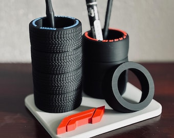May include: A desk organizer set featuring two black tire-shaped pen holders, one loose tire, and a red F1 logo emblem on a white square base. The taller tire holder has a blue rim detail, while the shorter one has red rim accents.