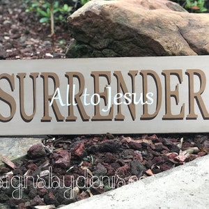 May include: A wooden sign with the words "SURRENDER All to Jesus" in brown lettering on a beige background.