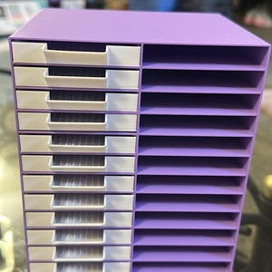 Puede incluir: Un organizador de escritorio morado con diez cajones deslizantes blancos en el lado izquierdo y diez ranuras de almacenamiento horizontales abiertas en el lado derecho. La unidad tiene un marco rectangular con una superficie superior plana.