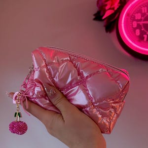 May include: A shiny, quilted pink pouch featuring a metallic finish and a zipper closure. Attached to the zipper pull is a decorative charm shaped like a pair of cherries, with green stems and two sparkling pink crystal-encrusted fruit spheres.