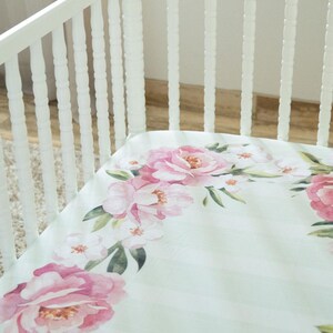 May include: A white crib with a floral patterned sheet. The sheet features a watercolor design of pink roses and green leaves on a light green striped background. The crib is made of white wood.