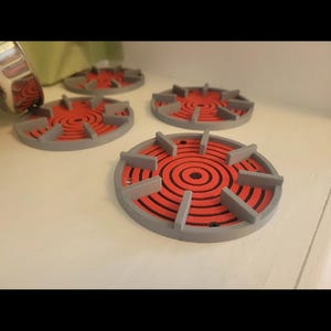 Könnte beinhalten: Drei kreisförmige Untersetzer mit einem grauen Außenring und erhöhten grauen Speichen, die ein leuchtend rotes Zentrum mit konzentrischen kreisförmigen Rillen umgeben. Das Design ahmt einen Herdplattenrost nach.