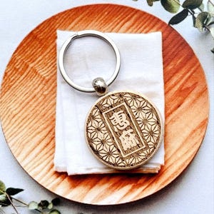 May include: A circular wooden keychain featuring a laser-engraved geometric star pattern surrounding a central rectangular frame. Inside the frame are two Japanese kanji characters. The keychain is attached to a standard silver-colored metal ring.