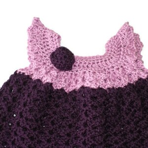 May include: A crocheted baby dress with a purple and pink color scheme. The dress has a flower-shaped embellishment on the front.