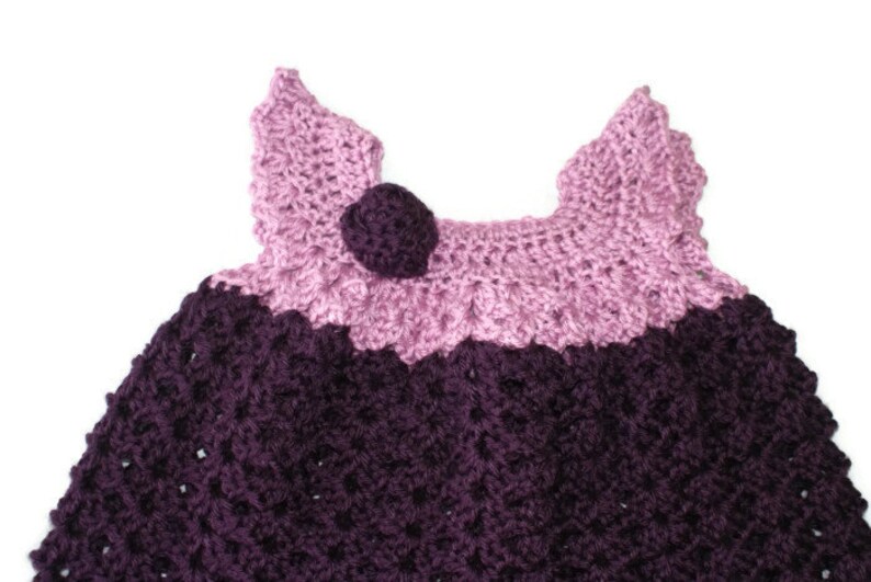 May include: A crocheted baby dress with a purple and pink color scheme. The dress has a flower-shaped embellishment on the front.