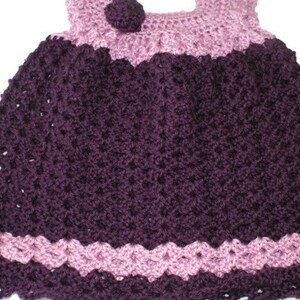 May include: A crocheted baby dress with a purple and pink color scheme. The dress has a scalloped hem and a small flower embellishment on the bodice.