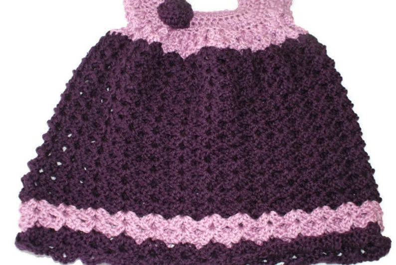 May include: A crocheted baby dress with a purple and pink color scheme. The dress has a scalloped hem and a small flower embellishment on the bodice.