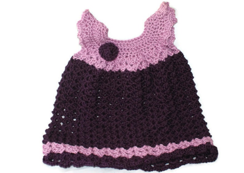 May include: A crocheted baby dress with a purple and pink color scheme. The dress has a flower embellishment on the bodice.