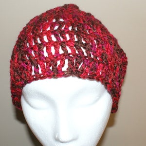 May include: A crocheted beanie hat in shades of red, brown, and pink. The hat is made with a loose, open stitch pattern.