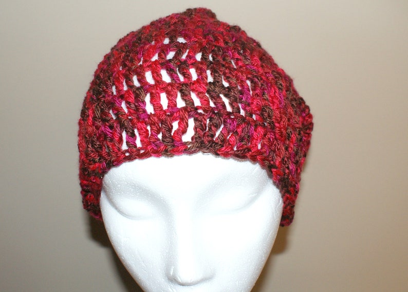 May include: A crocheted beanie hat in shades of red, brown, and pink. The hat is made with a loose, open stitch pattern.