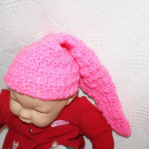 May include: A pink crocheted baby hat with a long pointed tail. The hat is worn by a baby doll with a red shirt.
