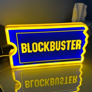 Peut inclure: Une enseigne LED rectangulaire en forme de ticket avec une bordure jaune vif et un centre bleu profond. Le mot BLOCKBUSTER est affiché en lettres capitales jaunes, grasses et sans empattement au milieu de l'enseigne lumineuse.