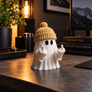 May include: A white ghost figurine wearing a tan knit beanie with a pom-pom on top. The ghost has two black oval eyes and is positioned with both arms raised, extending its middle fingers in a gesture. The figure has a smooth, matte finish.